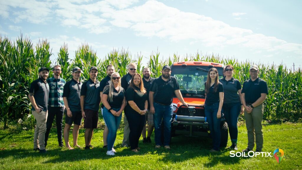 The SoilOptix® team standing in front of a field of tall corn beside an orange field vehicle, showcasing the people behind high-resolution soil mapping services.